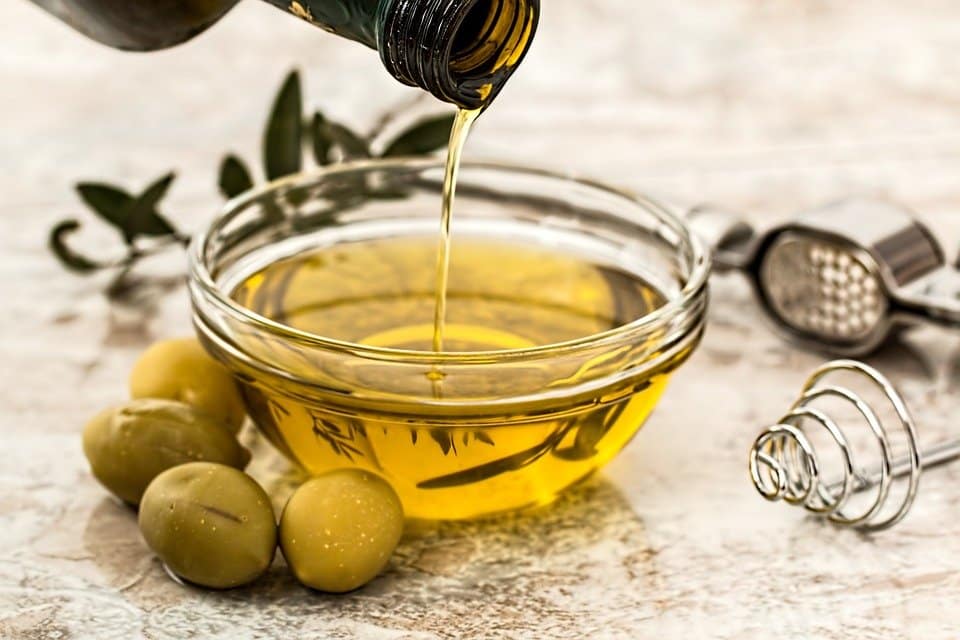 Olive oil being poured into small glass bowl with olives around it