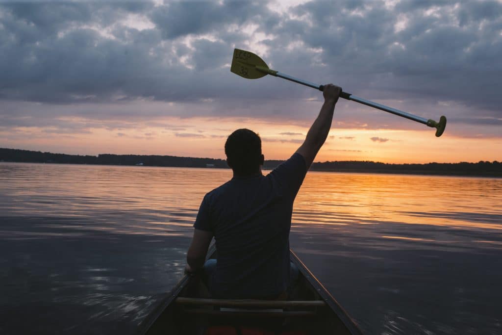Person in boat with oar in hand and hand stretched out above head in the sunset